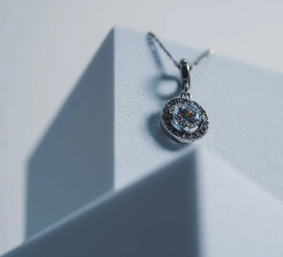 Round halo diamond pendant on a fine chain, displayed on a light stand