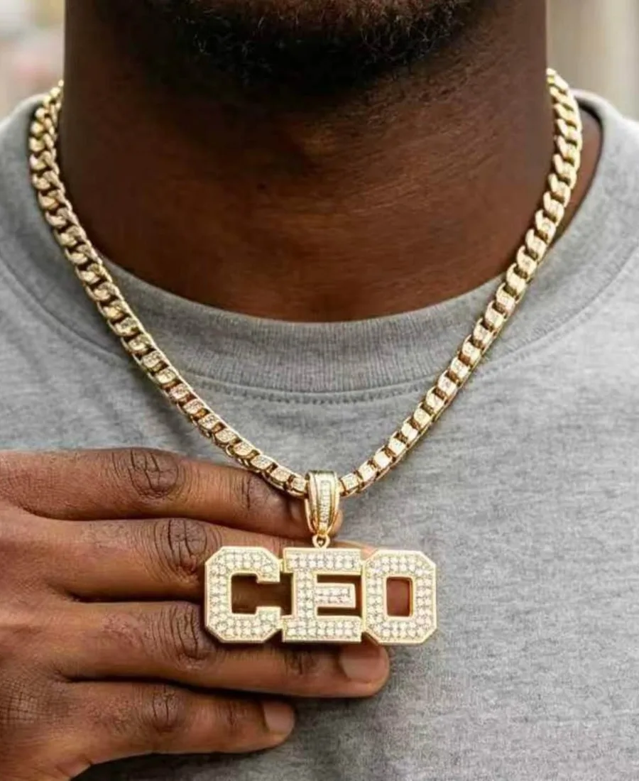 Man in a grey tee holding an iced block-letter pendant on a gold Cuban chain
